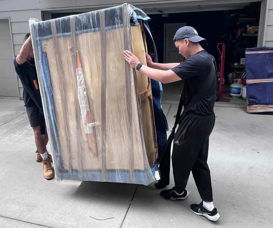 Movers carrying a shrink-wrapped dresser out of a garage